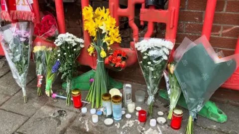 BBC Several bunches of flowers are lined up along a red brick wall. On the ground are several red and white candles. 