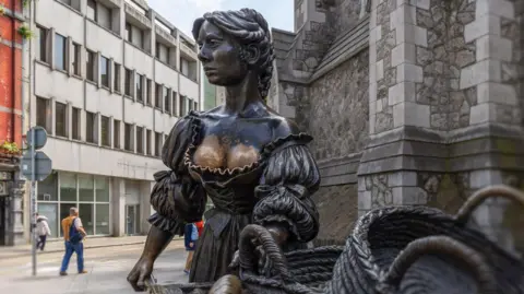 Artem Bolshakov/Getty Images The bronze statue of Molly Malone in St Andrew's Street in Dublin.  The artwork depicts a young woman in historical dress holding a wheelbarrow full of baskets.  The outfit exposes part of the sculpture's breasts which now appear gold in colour rather than dark bronze.  