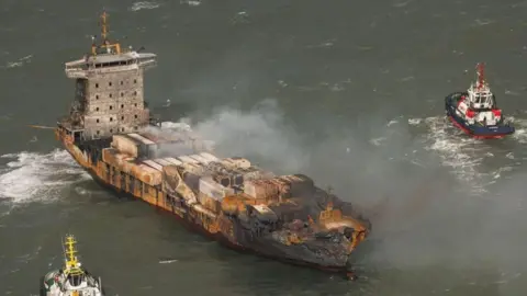 EPA-EFE/REX/Shutterstock The Solong cargo ship with a fire-damaged hull and smoke rising from the deck, with two tug boats nearby,