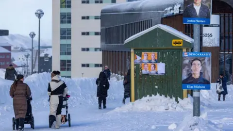 Getty Images Two women with pushchairs chat as they walk past a bus stop showing several election campaign posters, with snow on the ground all around