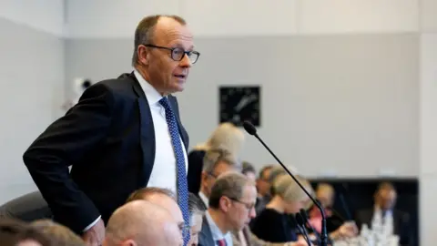 Reuters Friedrich Merz stands in front of a microphone wearing a suit and tie to talk to reporters