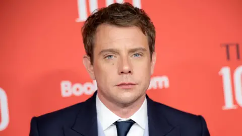 Getty Images Jonathan Anderson stands in front of a large, red wall and looks at the camera. He has short hair, a navy suit jacket, a white collared shirt and a navy tie.