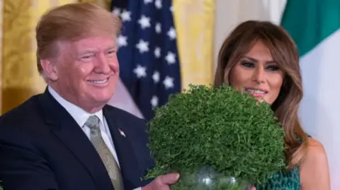 Getty Images President Donald Trump holding bowl of shamrock, he is smiling and wearing a dark blue suit and gold tie with a white shirt. Beside him is his wife Melania who is wearing a dark green dress.