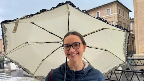 BBC A young woman wearing glasses stands under an umbrella