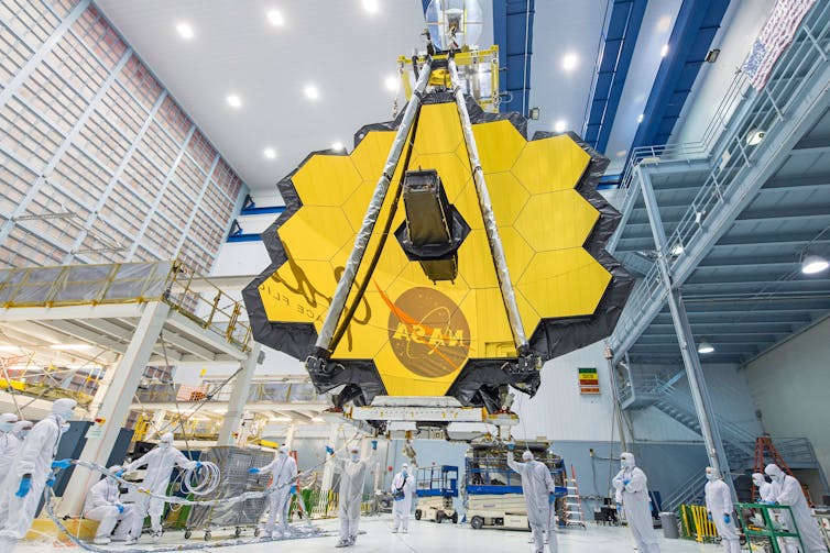 The JWST's mirror, which looks like a large, roughly hexagonal shiny surface made up of 18 smaller hexagons put together, sitting in a facility. The mirror is reflecting the NASA meatball logo.