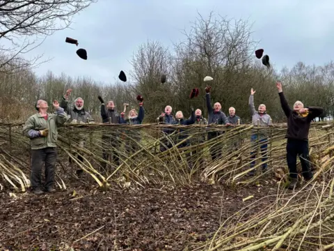 Earth Trust A group of volunteers stand around a hedge that they are laying. They are throwing their hats in the air and smiling.