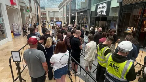 BBC Men and women are queued up near railings outside the Oasis Live '25 pop-up shop. Two men are wearing yellow high-visibility vests and one has a hard hat. A sign directs people where to queue. The Bullring shopping centre can be seen in the background.