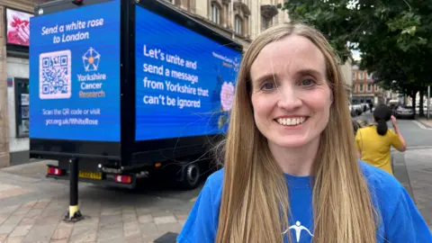 BBC News A woman with long blond hair wears a blue charity T-shirt and smiles as she stands next to a blue van displaying digital billboards for Yorkshire Cancer Research. Messages read: "Send a white rose to London", and "Let's unite and send a message from Yorkshire that can't be ignored". 