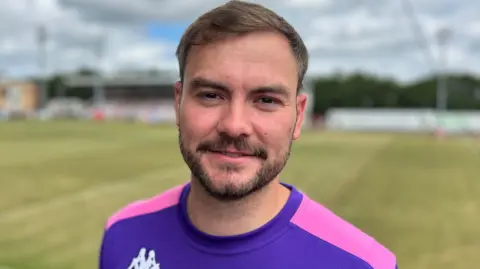 BBC 30-year-old man with brown hair and facial hair at the Batley Bulldog's rugby league stadium wearing a purple and pink rugby top smiling at the camera.