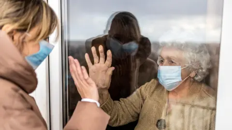 PIKSEL/Getty Images. A stock photo of an older woman waving at a younger woman through the window of a care home.  They are both wearing medical face masks. The older woman has short, curly white hair and is wearing a beige jumper.  The younger woman has blonde hair tied back and is wearing a light brown hooded coat. 