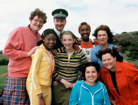 A colourful line up of chacters in tracksuits, kilts and jumpers ahanist a windy island background.