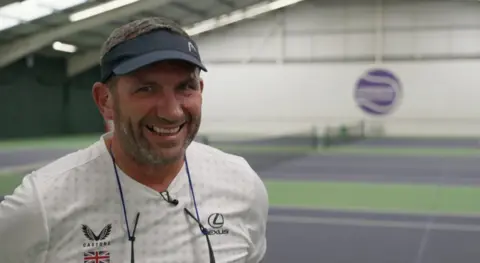 BBC Neil Bamford is standing on the tennis courts and he is staring directly at the camera and laughing. He is wearing a black visor and white tennis T-shirt. 