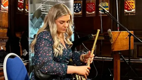 Handout A young woman with blond long wavy hair playing the uilleann pipes on a blue chair. Behind her are brown panels with emblems and a grey pop-up sign. She is looking down at the uilleann pipes.