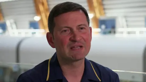 BBC A man sits speaking wearing a navy nurse's uniform in a hospital building.
