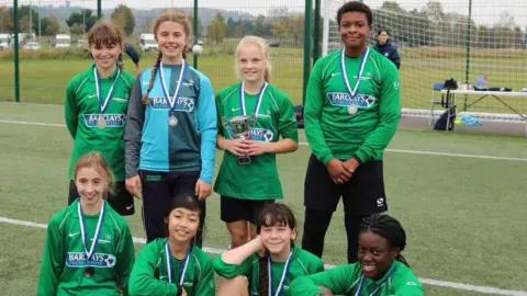 Supplied Two rows of smiling young footballers on a girls team, they're wearing green and have medals round their necks, a blonde girl on the top row is holding a trophy. 