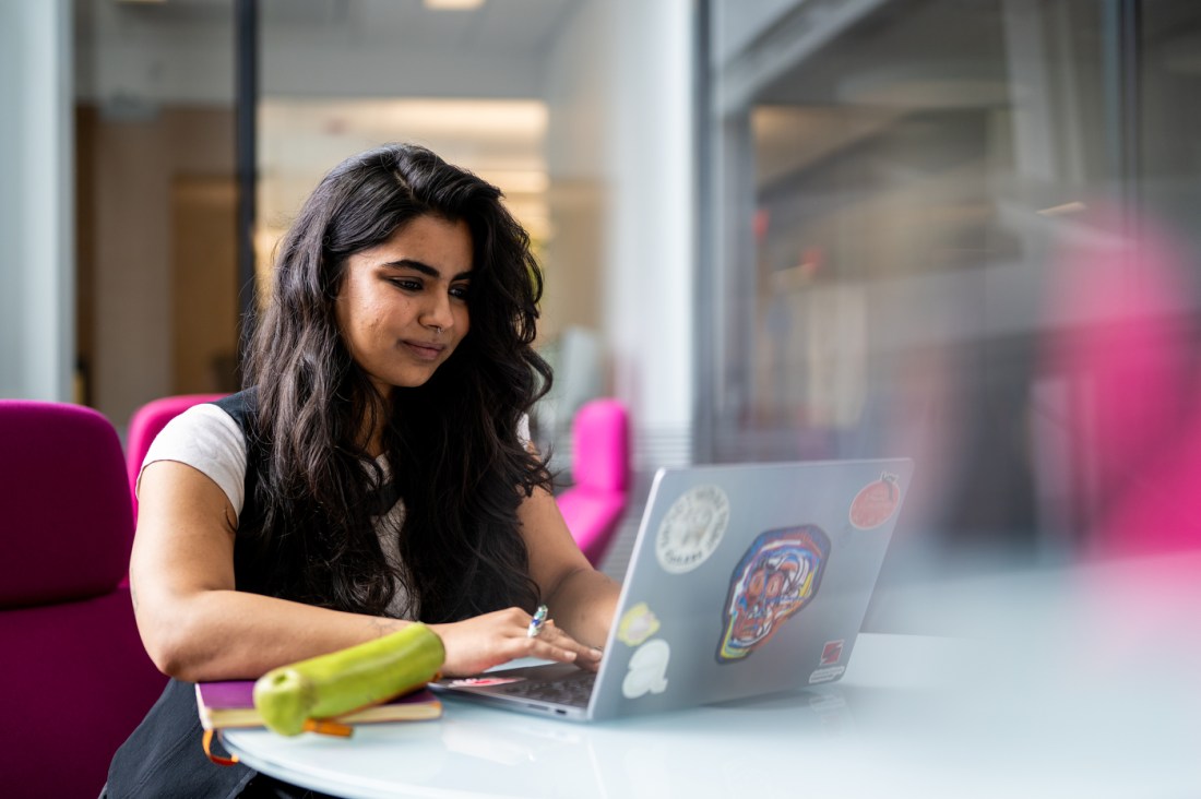 Mukki Gill sitting at a round white table working on her laptop, which is covered with an array of stickers.