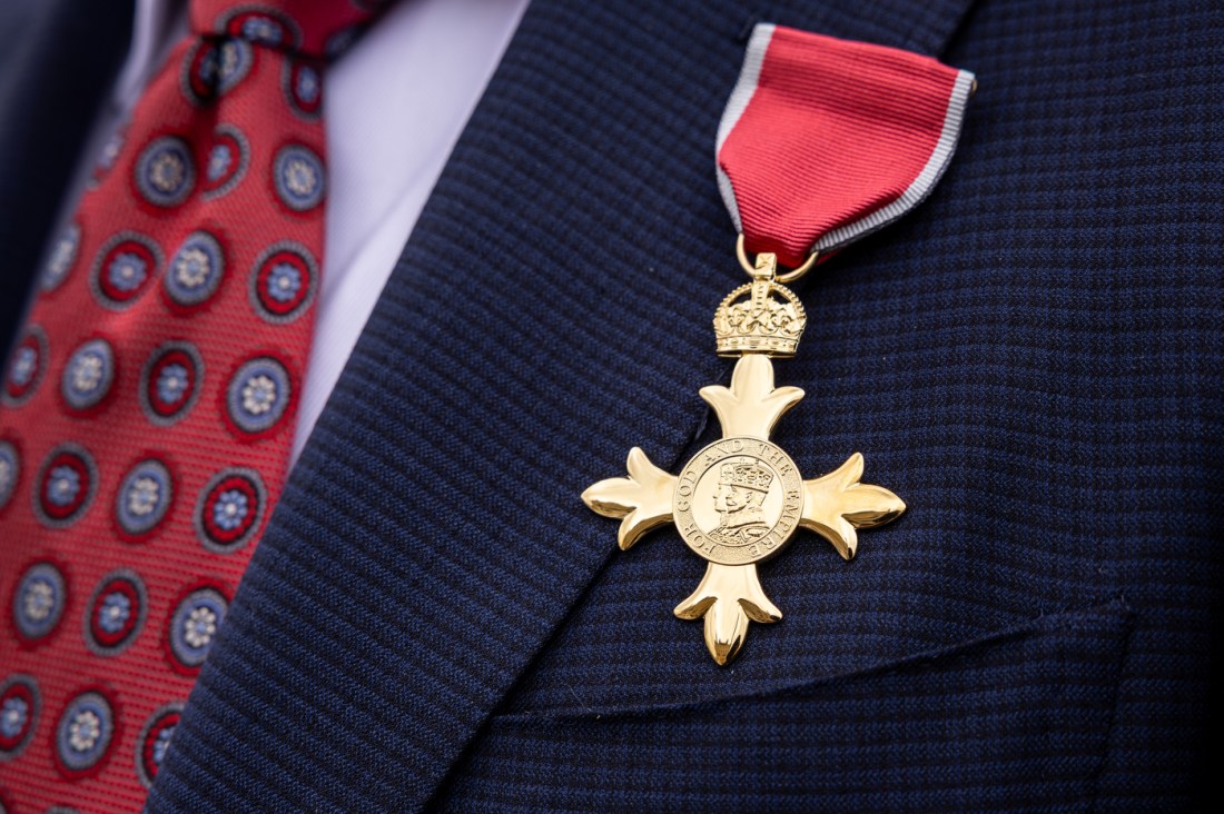 A close-up of a gold metal with a red ribbon on a dark blue suit jacket next to a red tie.