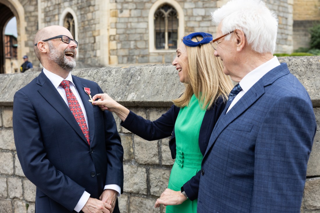 A bearded man in a dark suit and red tie having a medal adjusted on his lapel by a women in a bright green dress. They are laughing. Next to the woman is a man with white hair and glasses watching and also smiling.