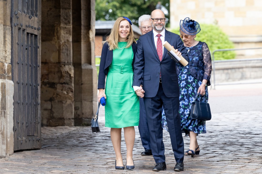 Ken Henderson and his wife and parents stand together on cobblestones outside a historic stone building. Ken Henderson is wearing a navy suit and red tie, his wife is wearing a bright green dress and navy blazer, and his mother is wearing a blue floral dress and matching hat. His father stands behind them in the background.