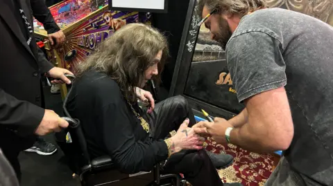 Luke McDonnell Ozzy Osbourne, in a wheelchair, is shaking hands with a man who is stood up. The man is holding a yellow marker pen.