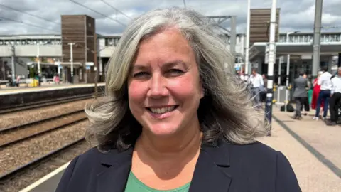 Ben Schofield/BBC Heidi Alexander wearing a black blazer over a green T-shirt. She is smiling while standing on a train platform.