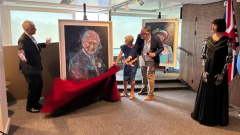 Copyright 2025 © Ai-Da Robot Studios The portrait of King Charles III unveiled at the United Nations' AI for Good Global Summit. Ai-Da is standing to the side while her inventor Aidan Meller and two other speakers pull the red veil down to showcase the portrait. Ai-Da's portrait of the late Queen Elizabeth II is behind them.