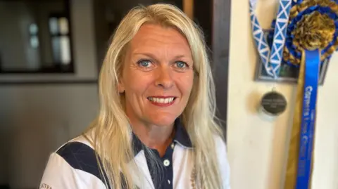 A woman with long, blonde hair in a blue and white T-Shirt with medals and rosettes in the background