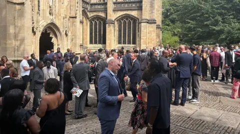 Dozens of people stand outside St Mary Redcliffe Church in Bristol after the funeral for former Gloucestershire and England cricketer David "Syd" Lawrence. They are mostly in dark suits or dresses
