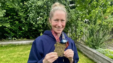Anna Rutherford Anna Rutherford looking right at the camera and smiling. She is holding a large gold medal in front of her body. She has fair hair and is wearing a blue hooded top. She is standing in a garden surrounded by green plants and grass.