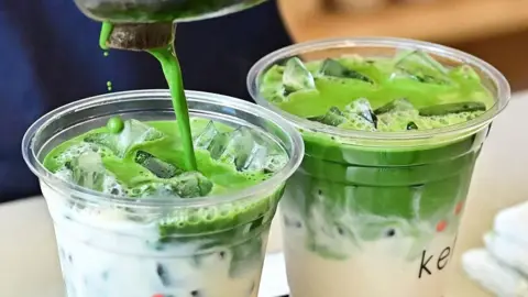 Getty Images The photo shows two cups of matcha latte being prepared, with the bright green tea being poured into one of the plastic cups filled with milk. 