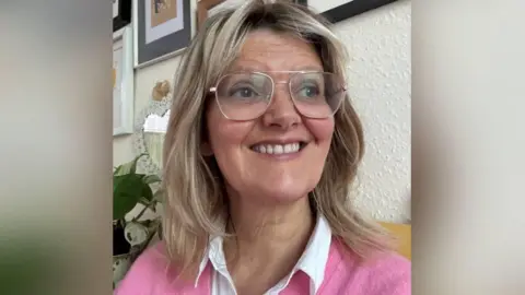 Iola Ynyr Iola Ynyr is smiling as she looks just away from the camera. She has long blonde hair and is wearing glasses, a white blouse and a pink jumper.