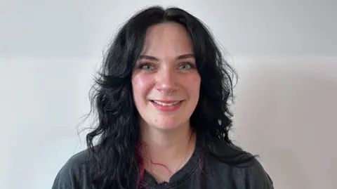 Beth Parsons/BBC Abbey Parrinello, who has black hair with red tips and blue eyes. She has a septum piercing and is smiling in the photo.