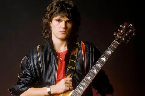 Getty Images A young Robin George wears a black leather jacket and red t-shirt with a guitar slung over his left shoulder.