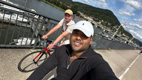 Ziyad Azad Ziyad Azad and Aaron Moloney cycling along the waterfront with boats and a large hill behind them. Mr Azad is taking the selfie photo while cycling and wearing a white hat and black fleece. Mr Moloney is seen in the background wearing a beige cap and an off-white polo top. 