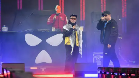 Getty Images Two rappers perform on stage with microphones, wearing sunglasses and streetwear, while a masked DJ stands behind a booth decorated with a white skull-like symbol. Purple and blue lights fill the stage.
