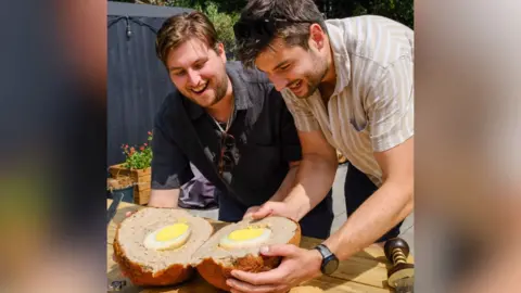 Oli Paterson and Phoenix Ross Oli and Phoenix are holding their massive scotch egg, which is on a wooden table, that's been sliced open to reveal a giant ostrich egg baked inside. They are looking down at their creation and smiling.