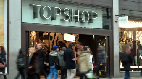 PA Media a bustling high street scene with a Topshop store as its centrepiece. Blurred shoppers are in the foreground, while the store front is brightly illuminated. large photos of models wearing the brand's clothing are seen in the windows.