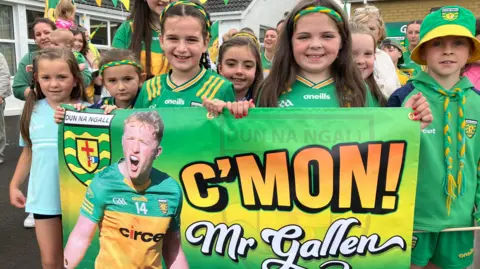 A group of girls in Donegal GAA gear hold up a green banner with a photo of teacher and Donegal player Oisín Gallen, who is in his Donegal county kit and is shouting. The banner says "C'Mon Mr Gallen" on it and it is green and yellow.