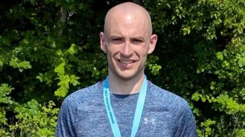 Thomas Mills Thomas Mills wears a blue top with a blue ribbon of a medal around his neck. The ribbon says "HALF DRAM 2025 FINISHER". He smiles and stands in front of a tree with lots of green leaves.