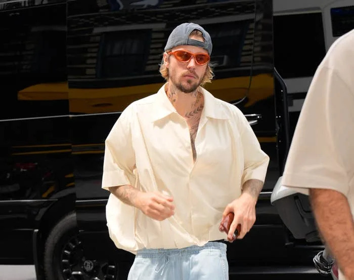 A person in a casual oversized shirt, light pants, and hat with sunglasses, walking outdoors in front of a vehicle
