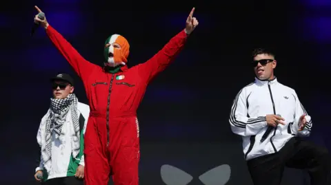 EPA/Shutterstock Kneecap on stage in outfits of white and red. Members to the left and right wearing shades and white clothing. Person in centre in red wearing balaclava of green white and orange.