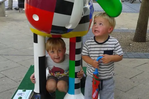 Family copyright A young Leo and Hugh crouch beneath a large, colourful sculpture during the 2015 trail. The sculpture is covered in bright designs and patterns. The boys smile beneath its large, floppy ears.