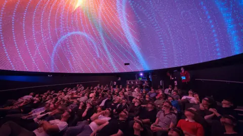Lee Pullen An audience looking up at the ceiling in a planetarium. The display is pink and there are blue dots all over it.