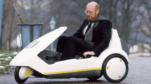 David Levenson Sir Clive Sinclair is sitting in a small electric vehicle in a photograph from 1985. He is mostly balding with auburn beard and moustache. He is wearing spectacles, a dark blue suit and a grey scarf. The three-wheeled vehicle is low to the ground and is white with black and yellow trims. There is a light dusting of snow on the grass in the background