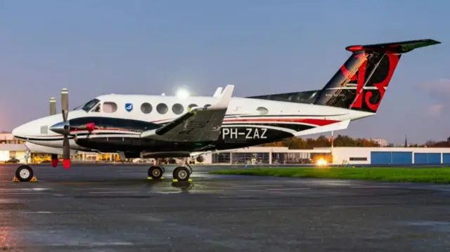 A small passenger plane. It is mostly white with black and red livery on its underside, engines and tail. It has one engine per wing. There are five round windows down the side behind the cockpit. It has "PH-ZAZ" on the fuselage.