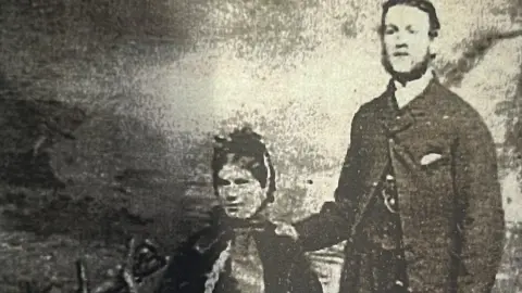 HANDOUT A faded black and white photo of a smartly dressed man and woman. She is sitting down and he stands with one hand on her shoulder. They are dressed in Victorian era clothing.