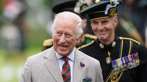 Reuters King Charles III wearing a grey suit with a white shirt and red and green striped tie. He is walking through the Palace Guard - a line of soldiers, wearing black uniforms.