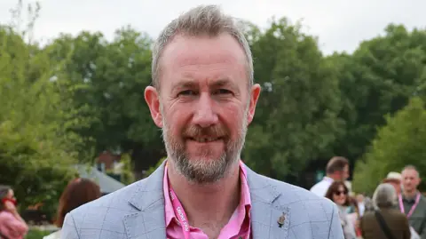 Getty Images A picture of Alex Horne, who is looking at the camera, smiling. He is wearing a pink shirt with a grey jacket and has people stood behind him.