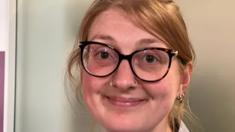 A close up if Serafina's face. She has tied back ginger hair and is wearing dark rimmed glasses and is smiling at the camera.