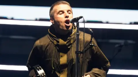 PA Media Liam Gallagher with tambourine and khaki jacket in Cardiff, He is singing into a microphone and has short dark hair.